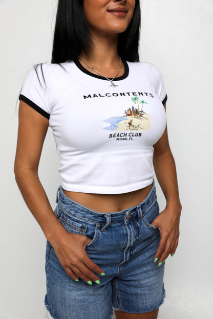 A white crop top t-shirt with black trim featuring a beach scene and text that reads "Malcontents Beach Club Miami FL.