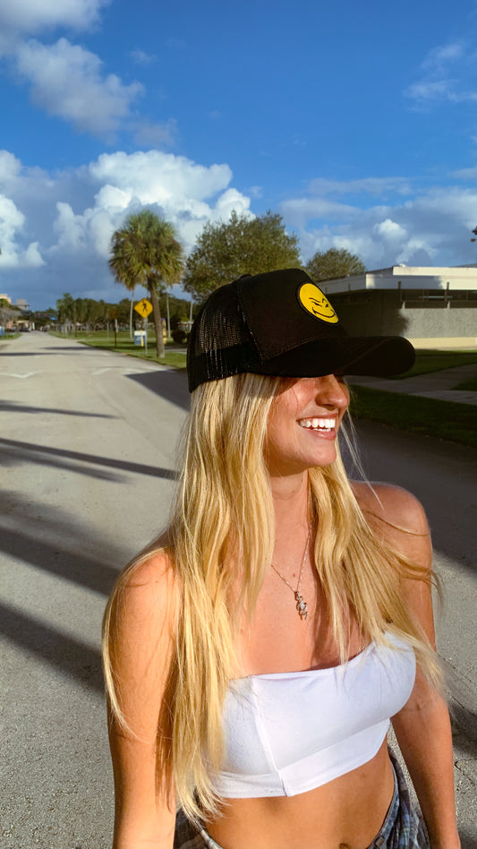 Malcontents Smiley Trucker Hat: Black trucker, mesh back, Malcontents smiley on front, Malcontents logo on side on blonde girl at the beach