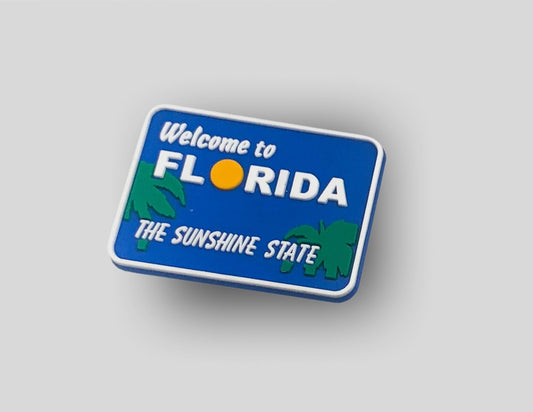 Malcontents Welcome to Florida Croc Charm/Jibbitz shows the blue "Welcome to Florida" sign with green palm trees, a yellow sun for the "O", and "The Sunshine State" in white letters.