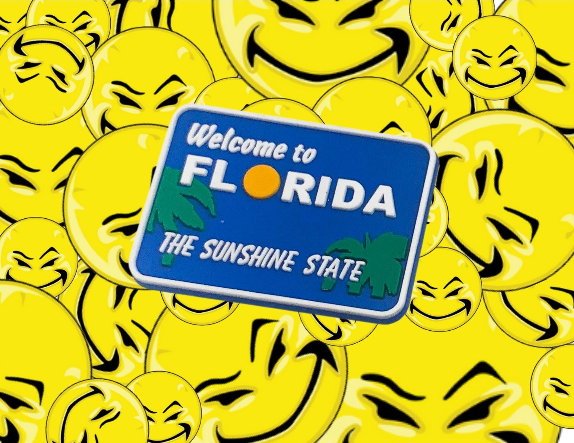 Malcontents Welcome to Florida Croc Charm/Jibbitz shows the blue "Welcome to Florida" sign with green palm trees, a yellow sun for the "O", and "The Sunshine State" in white letters in front of Malcontents smilies background